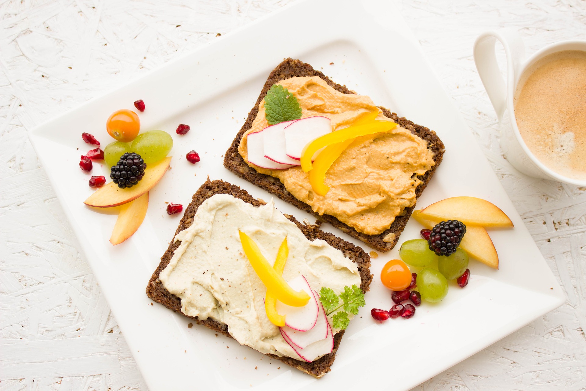 Fette di pane nero con hummus e crema chiara, ravanelli, peperone giallo, frutta fresca e cappuccino su piatto bianco